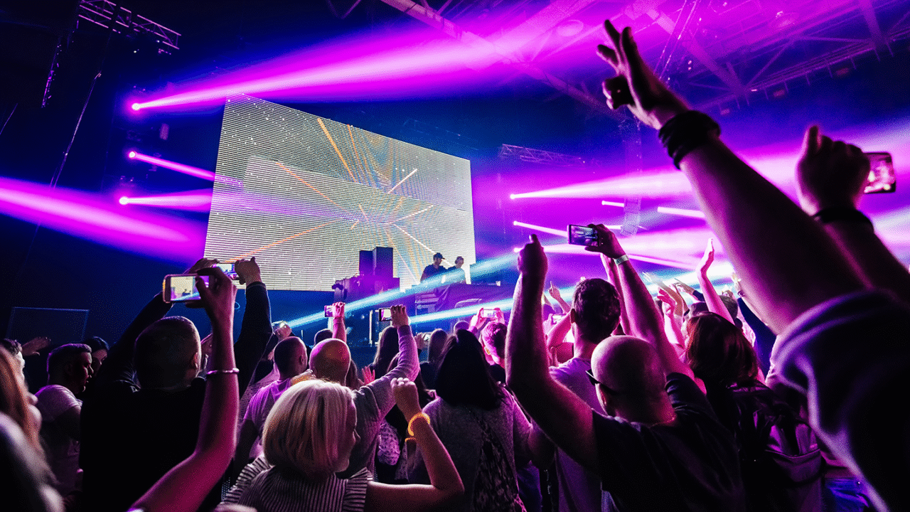 DJ performing on stage using effects with vibrant pink and purple lighting while the crowd raises their hands