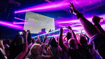 DJ performing on stage using effects with vibrant pink and purple lighting while the crowd raises their hands