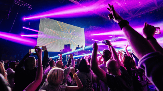 DJ performing on stage using effects with vibrant pink and purple lighting while the crowd raises their hands