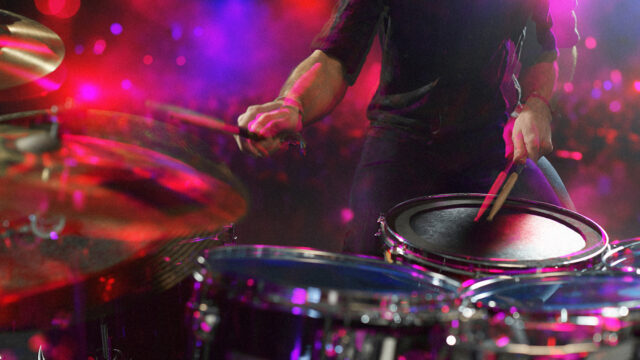 Drummer performing an energetic drum fill on stage with colorful lights during a live concert