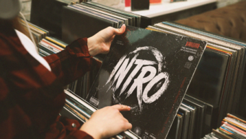 Person browsing vinyl records while holding a record sleeve reading "Intro", representing the best and most iconic song intros in music history.