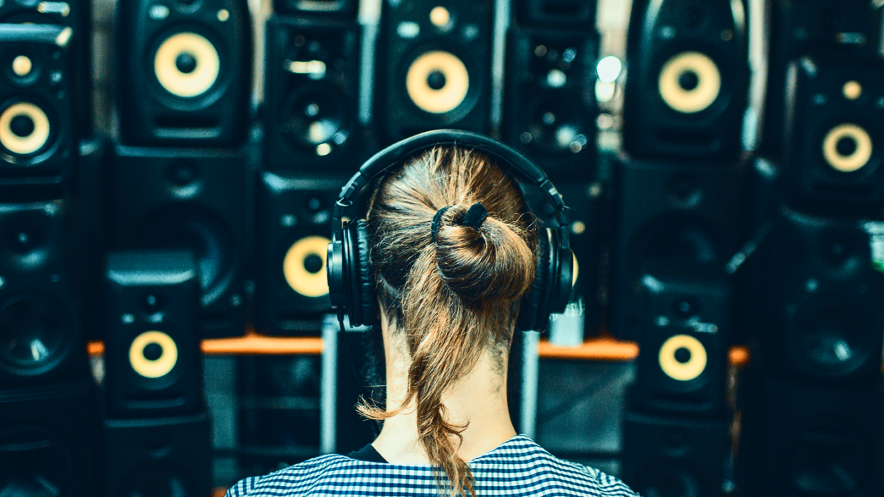 Person wearing headphones in front of a wall of studio speakers, illustrating the choice between studio monitors and headphones for music production.